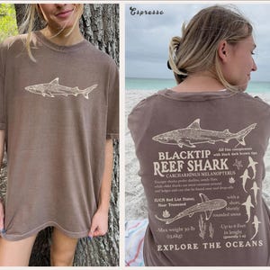 May include: Espresso-coloured t-shirt with a shark graphic on the front and back. The back of the shirt has text about the Blacktip Reef Shark, its features, and habitat. The shirt also includes the text "Explore the Oceans".