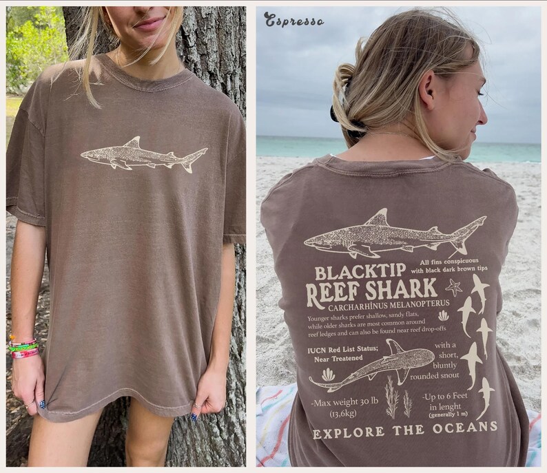 May include: Espresso-coloured t-shirt with a shark graphic on the front and back. The back of the shirt has text about the Blacktip Reef Shark, its features, and habitat. The shirt also includes the text "Explore the Oceans".