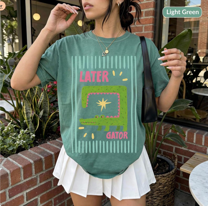 May include: Light green t-shirt with a cartoon alligator design. The design includes the words "LATER" and "GATOR" in pink and green, with a star and vertical stripes. The shirt is a casual style.