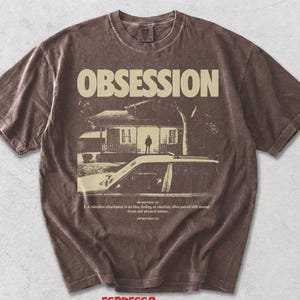 May include: A brown short-sleeve t-shirt featuring the word OBSESSION in large cream letters above a graphic of a house and a car. Below the graphic, the text reads: ob-ses-sion (n) 1. A relentless attachment to an idea, feeling, or emotion.