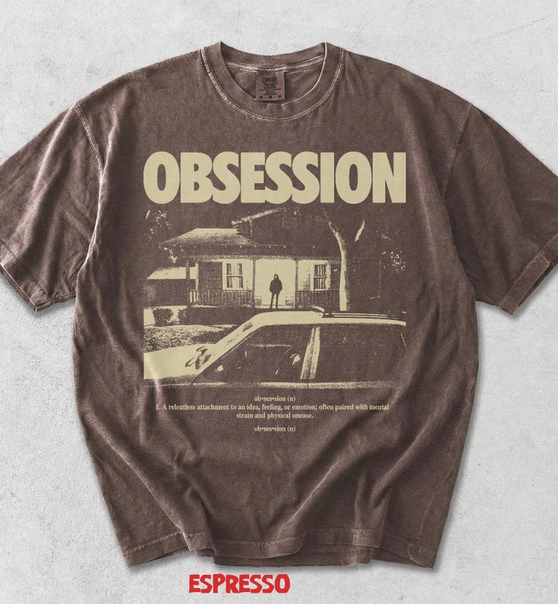 May include: A brown short-sleeve t-shirt featuring the word OBSESSION in large cream letters above a graphic of a house and a car. Below the graphic, the text reads: ob-ses-sion (n) 1. A relentless attachment to an idea, feeling, or emotion.