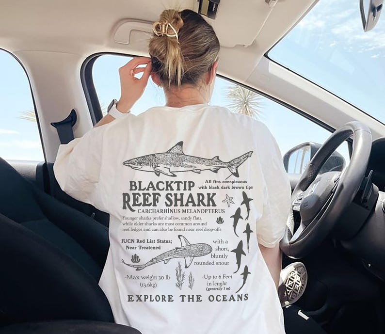 May include: Cream-coloured t-shirt featuring a detailed black and white illustration of a Blacktip Reef Shark, with accompanying text. The shirt includes scientific details and the words "EXPLORE THE OCEANS".