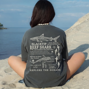 May include: Dark grey t-shirt with a detailed illustration of a Blacktip Reef Shark, and the text "BLACKTIP REEF SHARK" and scientific information. The shirt also includes the phrase "EXPLORE THE OCEANS".