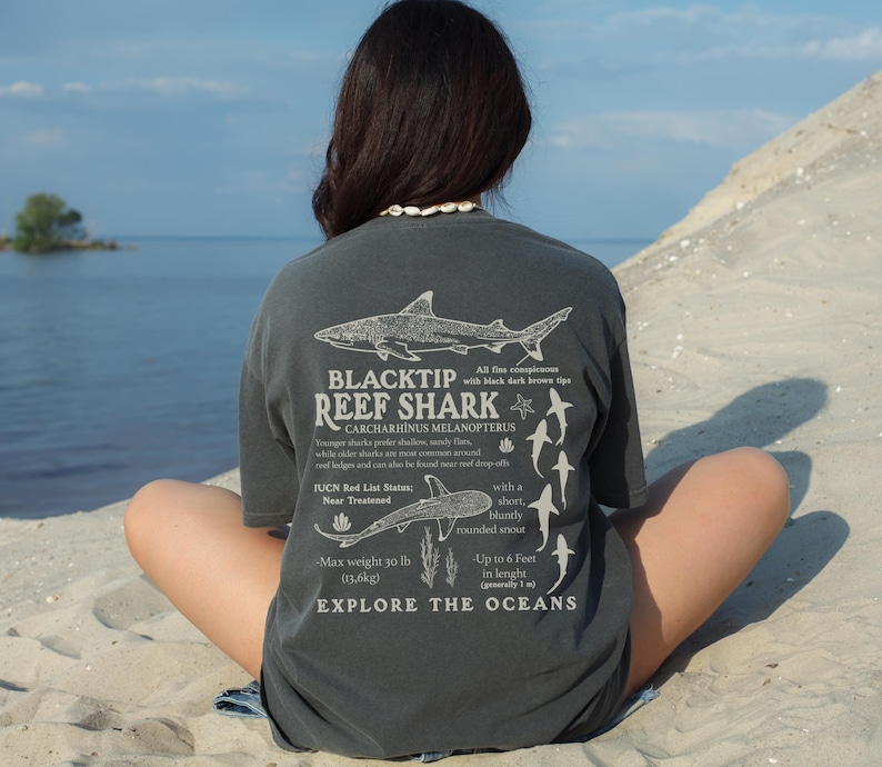 May include: Dark grey t-shirt with a detailed illustration of a Blacktip Reef Shark, and the text "BLACKTIP REEF SHARK" and scientific information. The shirt also includes the phrase "EXPLORE THE OCEANS".