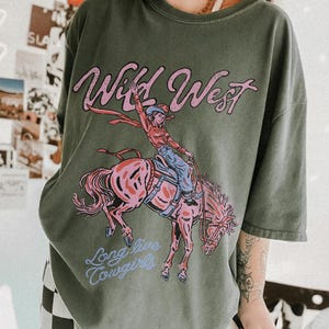 Puede incluir: Una camiseta verde oliva con un estampado gr&aacute;fico de una persona montando un caballo encabritado. El texto "Wild West" aparece en rosa sobre el jinete, y el texto "Long live cowgirls" aparece en azul claro bajo la ilustraci&oacute;n del caballo.