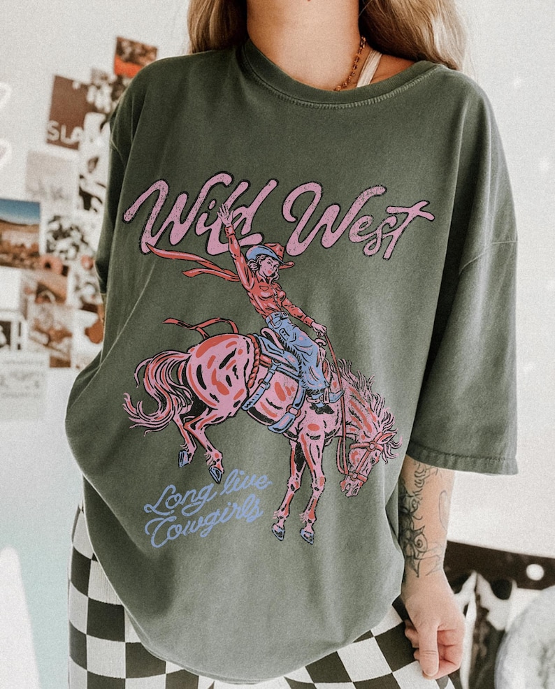 Puede incluir: Una camiseta verde oliva con un estampado gr&aacute;fico de una persona montando un caballo encabritado. El texto "Wild West" aparece en rosa sobre el jinete, y el texto "Long live cowgirls" aparece en azul claro bajo la ilustraci&oacute;n del caballo.