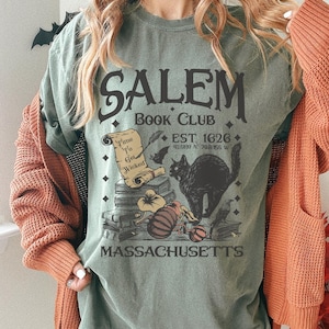May include: Sage green t-shirt with a Salem Book Club graphic. The design includes a black cat, pumpkins, books, and the text "Time To Get Wicked." The shirt also features the text "EST. 1626" and "MASSACHUSETTS."