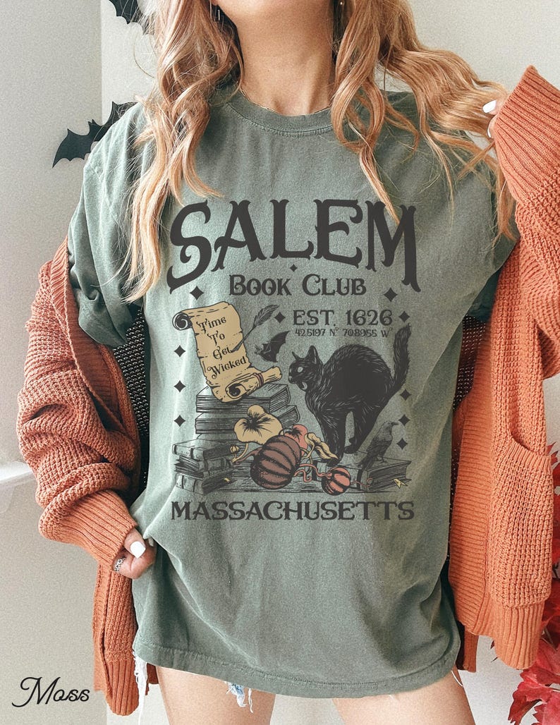 May include: Sage green t-shirt with a Salem Book Club graphic. The design includes a black cat, pumpkins, books, and the text "Time To Get Wicked." The shirt also features the text "EST. 1626" and "MASSACHUSETTS."