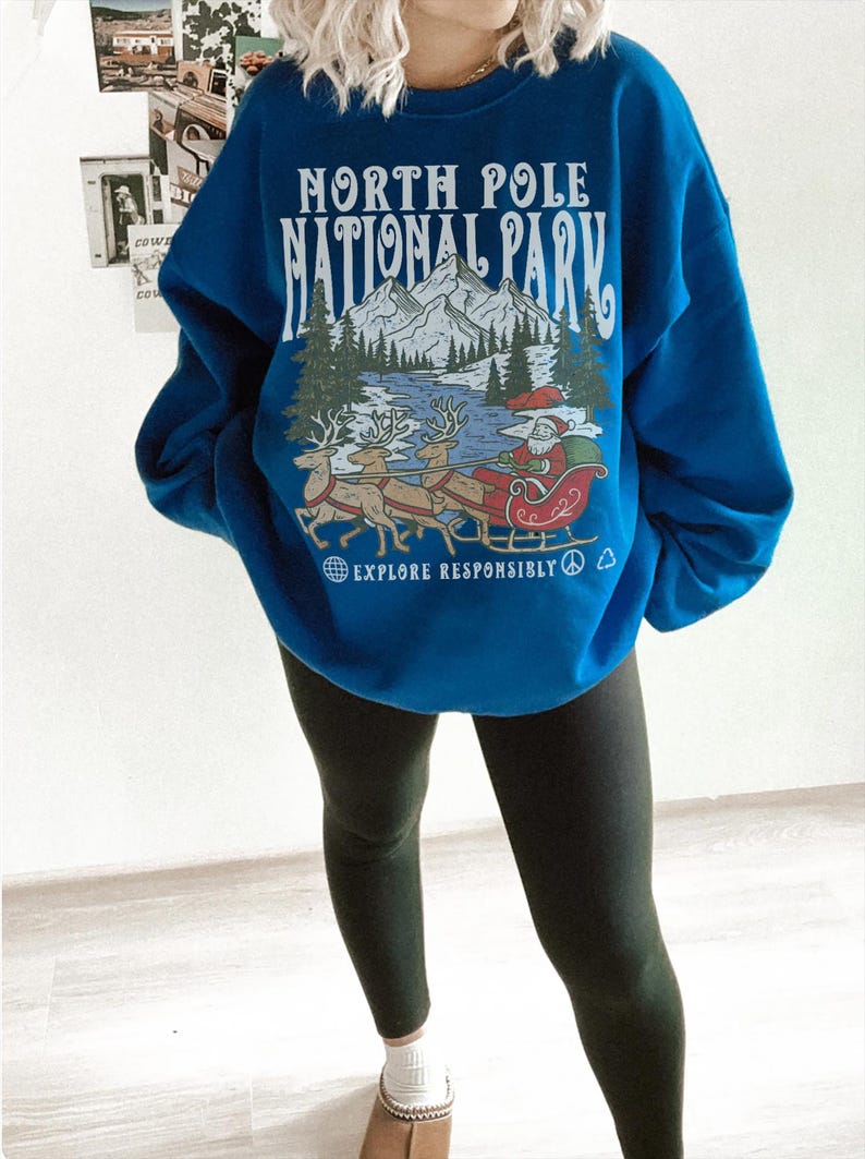 May include: A blue sweatshirt with the text "NORTH POLE NATIONAL PARK" above a graphic of Santa Claus in a sleigh pulled by reindeer. The phrase "EXPLORE RESPONSIBLY" is below the graphic. The person is wearing black leggings.