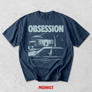May include: A dark blue t-shirt featuring a light blue graphic print of a house at night with a car in the foreground. The text reads OBSESSION at the top and includes a dictionary-style definition below the image. The word MIDNIGT appears in red.