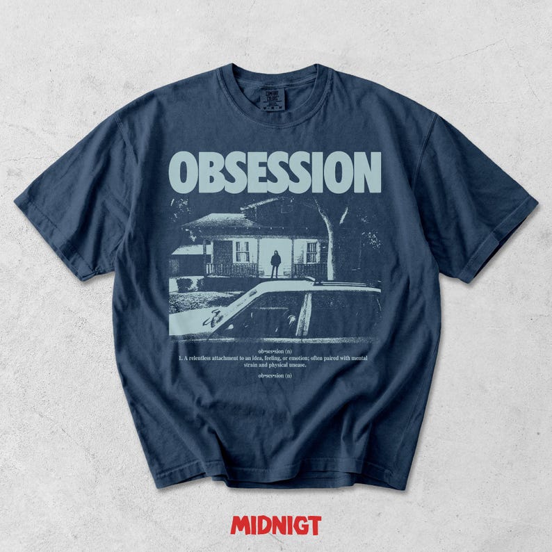 May include: A dark blue t-shirt featuring a light blue graphic print of a house at night with a car in the foreground. The text reads OBSESSION at the top and includes a dictionary-style definition below the image. The word MIDNIGT appears in red.