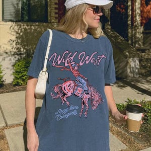 Puede incluir: Una camiseta azul pizarra con un estampado gr&aacute;fico de una vaquera montando un caballo rosa que corcovea. El texto "Wild West" aparece en rosa sobre la ilustraci&oacute;n, y las palabras "Long live cowgirls" est&aacute;n impresas en azul claro debajo.