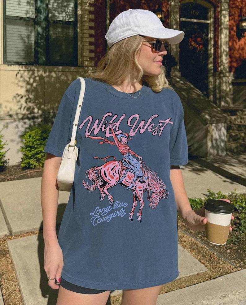 Puede incluir: Una camiseta azul pizarra con un estampado gr&aacute;fico de una vaquera montando un caballo rosa que corcovea. El texto "Wild West" aparece en rosa sobre la ilustraci&oacute;n, y las palabras "Long live cowgirls" est&aacute;n impresas en azul claro debajo.
