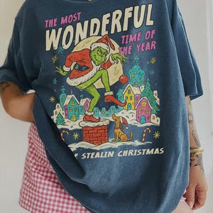 Puede incluir: Camiseta azul con un gr&aacute;fico del Grinch con gorro de Pap&aacute; Noel, con el texto "THE MOST WONDERFUL TIME OF THE YEAR" y "STEALIN CHRISTMAS". El Grinch est&aacute; sobre una chimenea. Un perro est&aacute; sentado cerca.