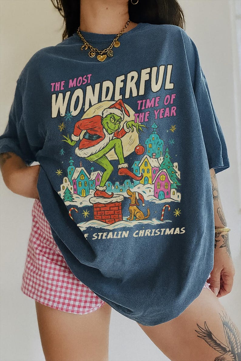 Puede incluir: Camiseta azul con un gr&aacute;fico del Grinch con gorro de Pap&aacute; Noel, con el texto "THE MOST WONDERFUL TIME OF THE YEAR" y "STEALIN CHRISTMAS". El Grinch est&aacute; sobre una chimenea. Un perro est&aacute; sentado cerca.