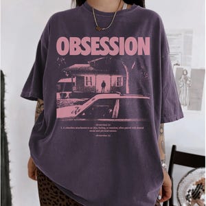 May include: A dark purple oversized t-shirt featuring the word OBSESSION in large pink letters above a graphic of a house and a car. Below the graphic, the text defines obsession as a relentless attachment to an idea, feeling, or emotion.