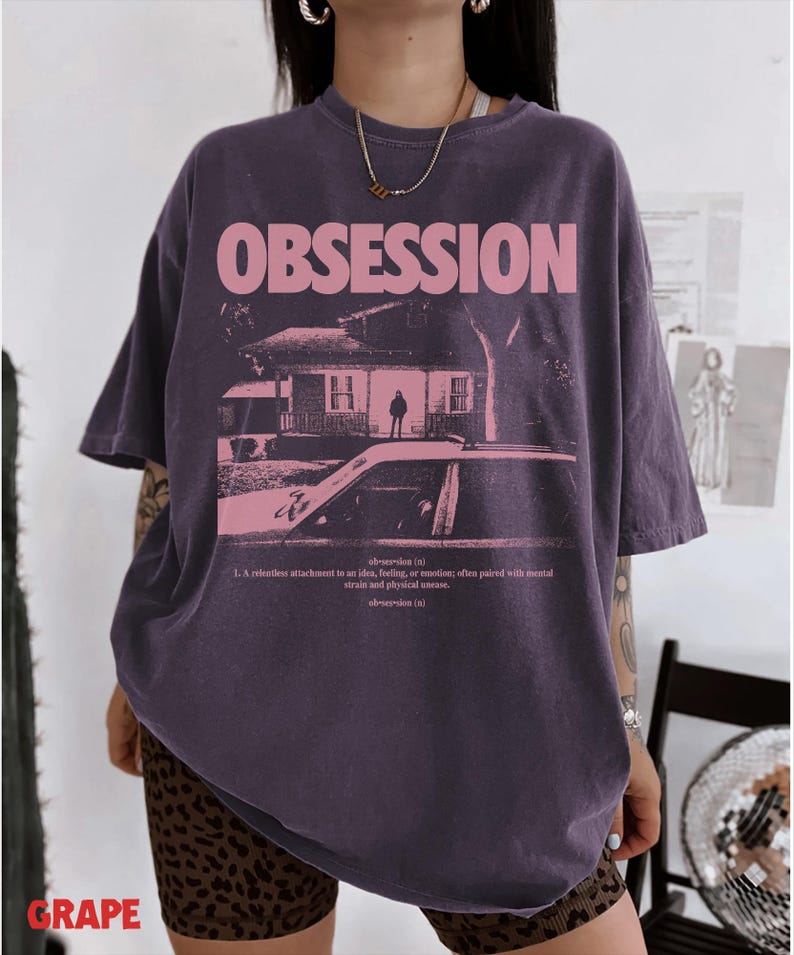 May include: A dark purple oversized t-shirt featuring the word OBSESSION in large pink letters above a graphic of a house and a car. Below the graphic, the text defines obsession as a relentless attachment to an idea, feeling, or emotion.