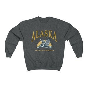 Peut inclure: Un sweat-shirt gris fonc&eacute; avec le texte "Alaska" en jaune, un soleil jaune et un aigle bleu volant au-dessus des montagnes. Le texte "The Last Frontier" est sous l'aigle.
