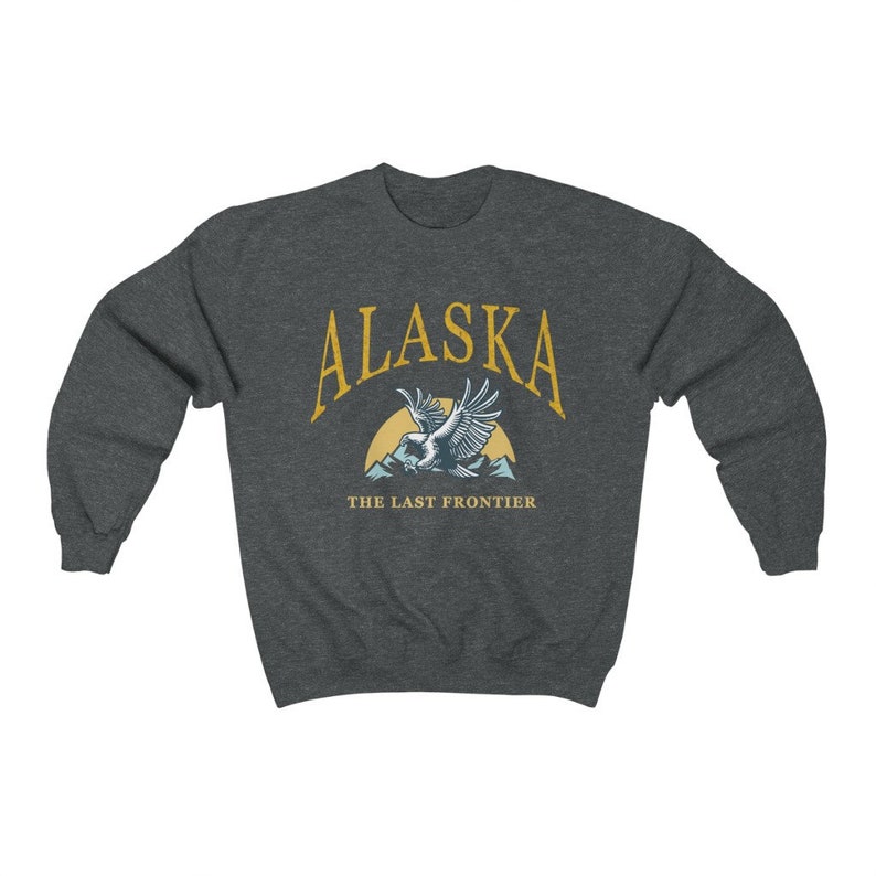 Peut inclure: Un sweat-shirt gris fonc&eacute; avec le texte "Alaska" en jaune, un soleil jaune et un aigle bleu volant au-dessus des montagnes. Le texte "The Last Frontier" est sous l'aigle.