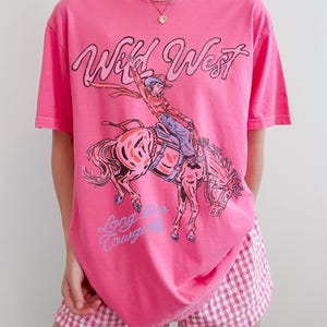 Puede incluir: Una camiseta rosa brillante de manga corta con un estampado gr&aacute;fico de una vaquera montando un caballo encabritado. El texto "Wild West" aparece en negro sobre la ilustraci&oacute;n y "Long Live Cowgirls" est&aacute; escrito en azul claro debajo.