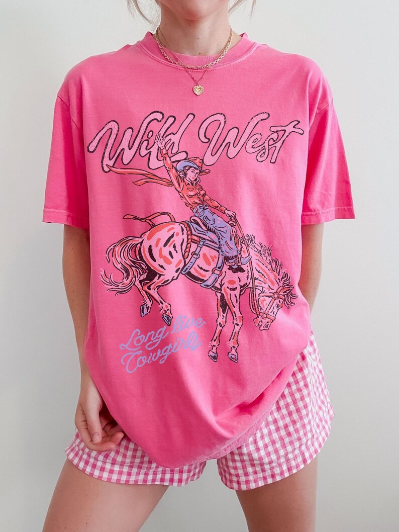 Puede incluir: Una camiseta rosa brillante de manga corta con un estampado gr&aacute;fico de una vaquera montando un caballo encabritado. El texto "Wild West" aparece en negro sobre la ilustraci&oacute;n y "Long Live Cowgirls" est&aacute; escrito en azul claro debajo.