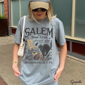 May include: A light blue t-shirt with the text "SALEM BOOK CLUB" and a graphic of a black cat, books, and a scroll. The shirt also includes the text "EST. 1626" and "MASSACHUSETTS".