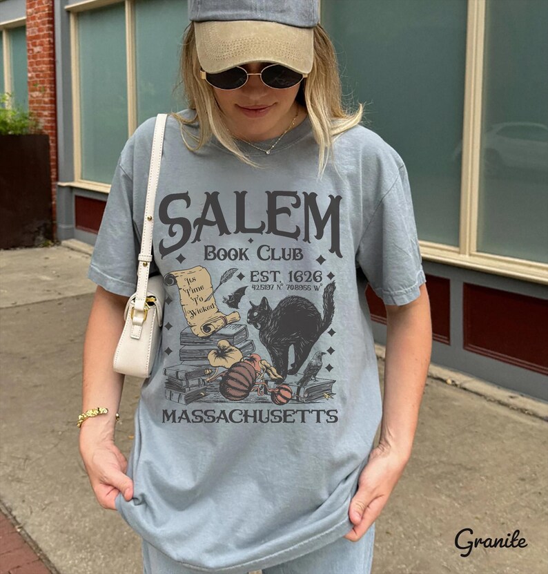 May include: A light blue t-shirt with the text "SALEM BOOK CLUB" and a graphic of a black cat, books, and a scroll. The shirt also includes the text "EST. 1626" and "MASSACHUSETTS".