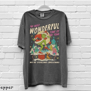 Puede incluir: Camiseta gris oscuro con un dise&ntilde;o navide&ntilde;o. El dise&ntilde;o incluye un personaje verde con gorro de Pap&aacute; Noel, un saco de regalos y el texto "The Most Wonderful Time of the Year" y "We're Stealing Christmas."