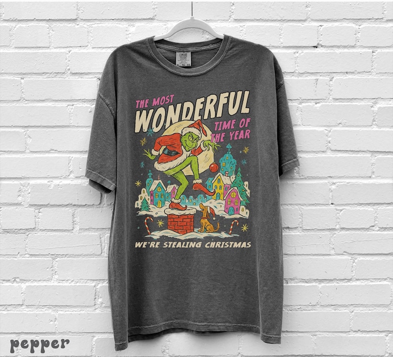 Puede incluir: Camiseta gris oscuro con un dise&ntilde;o navide&ntilde;o. El dise&ntilde;o incluye un personaje verde con gorro de Pap&aacute; Noel, un saco de regalos y el texto "The Most Wonderful Time of the Year" y "We're Stealing Christmas."