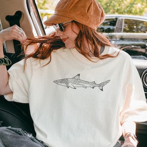 May include: Cream-coloured t-shirt with a detailed black and white illustration of a shark. The shark design is centered on the front of the shirt. The person in the image is wearing a brown corduroy cap and sunglasses.