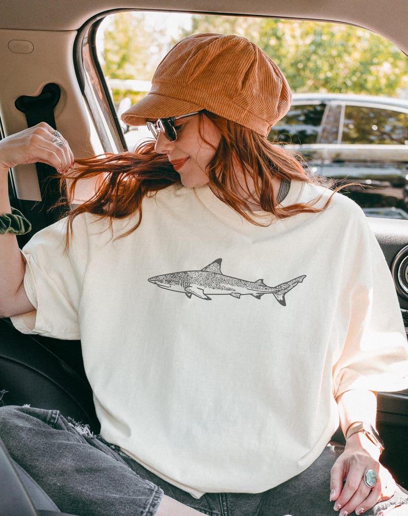 May include: Cream-coloured t-shirt with a detailed black and white illustration of a shark. The shark design is centered on the front of the shirt. The person in the image is wearing a brown corduroy cap and sunglasses.