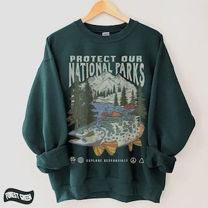 Puede incluir: Sudadera verde bosque con un gr&aacute;fico que dice "PROTECT OUR NATIONAL PARKS". El gr&aacute;fico presenta un pez, monta&ntilde;as, &aacute;rboles y el texto "EXPLORE RESPONSIBLY". La sudadera est&aacute; colgada en una percha de madera.