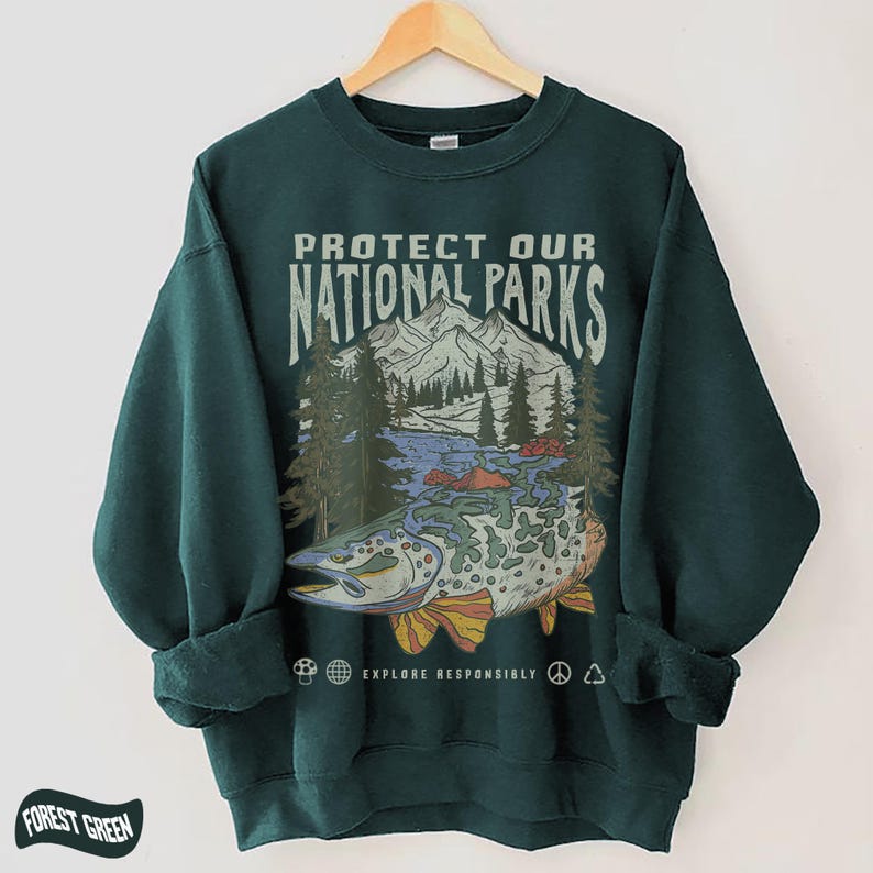 Puede incluir: Sudadera verde bosque con un gr&aacute;fico que dice "PROTECT OUR NATIONAL PARKS". El gr&aacute;fico presenta un pez, monta&ntilde;as, &aacute;rboles y el texto "EXPLORE RESPONSIBLY". La sudadera est&aacute; colgada en una percha de madera.