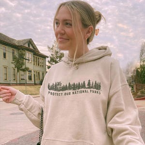 May include: A beige hooded sweatshirt featuring a dark green graphic print of a forest silhouette above the text "PROTECT OUR NATIONAL PARKS". The garment includes a front kangaroo pocket, long sleeves with ribbed cuffs, and a matching drawstring hood.