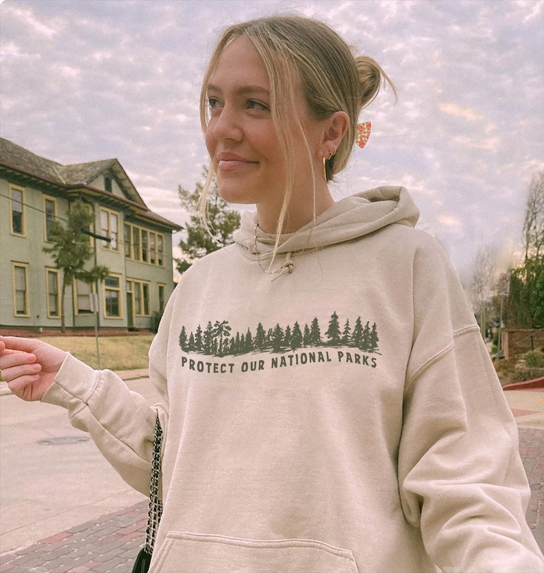 May include: A beige hooded sweatshirt featuring a dark green graphic print of a forest silhouette above the text "PROTECT OUR NATIONAL PARKS". The garment includes a front kangaroo pocket, long sleeves with ribbed cuffs, and a matching drawstring hood.