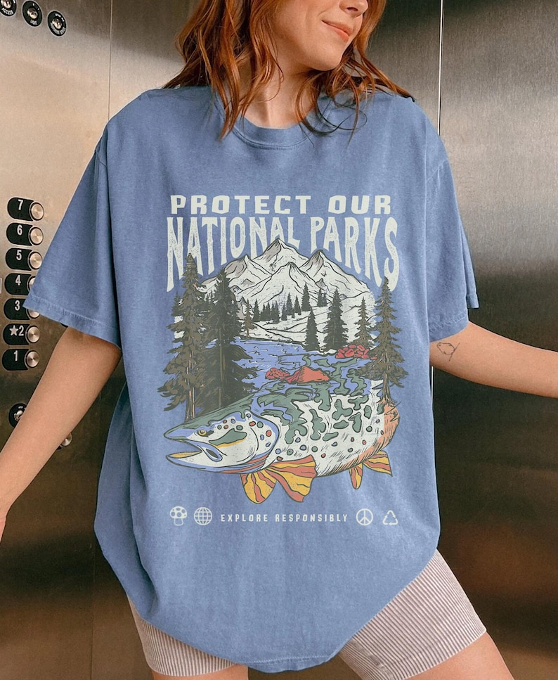 May include: A light blue t-shirt with a graphic print of a mountain scene and a fish. The text "PROTECT OUR NATIONAL PARKS" is printed on the shirt. The text "EXPLORE RESPONSIBLY" is printed below the graphic.
