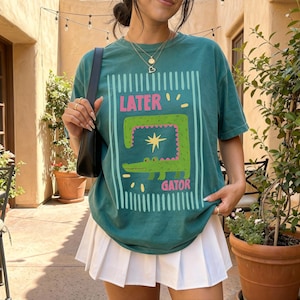 May include: Teal green t-shirt with a cartoon alligator design. The graphic includes the words "LATER" and "GATOR" in pink and white. The shirt is paired with a white pleated skirt.