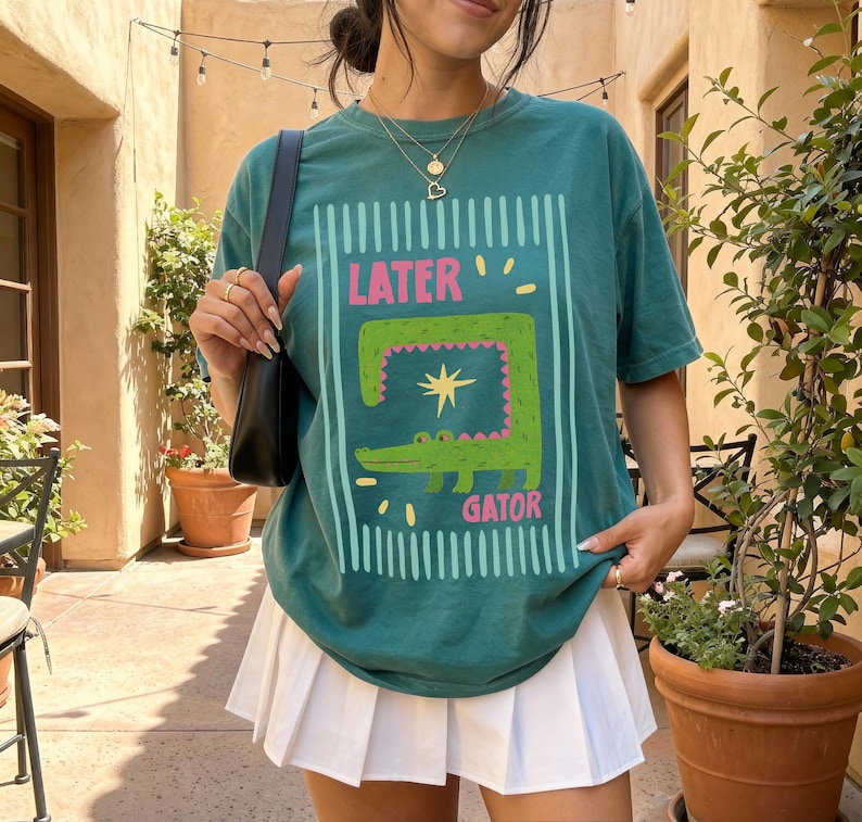 May include: Teal green t-shirt with a cartoon alligator design. The graphic includes the words "LATER" and "GATOR" in pink and white. The shirt is paired with a white pleated skirt.
