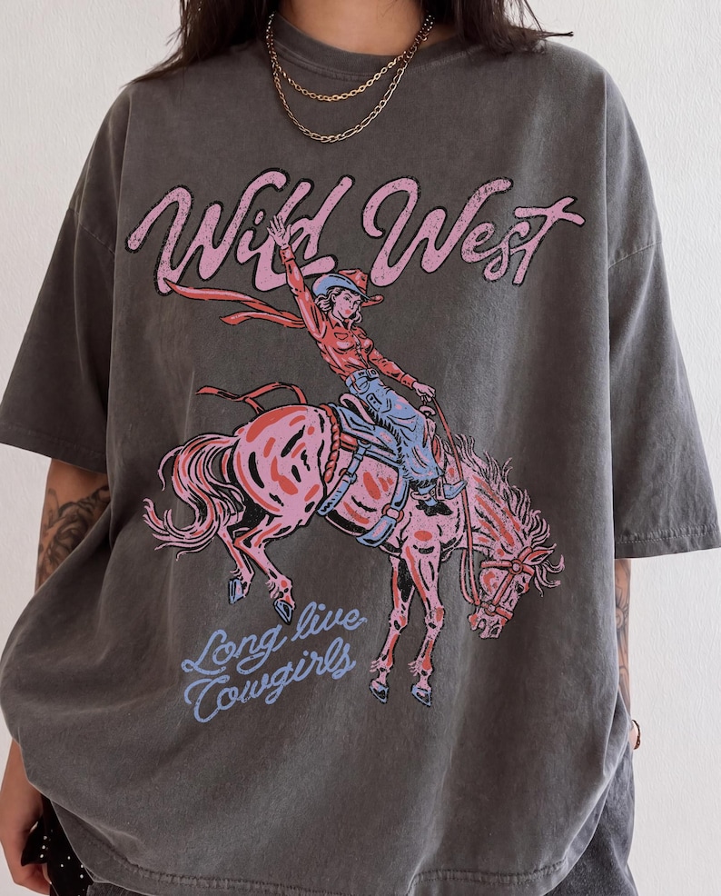 May include: A charcoal gray short-sleeve t-shirt featuring a graphic print of a person riding a bucking horse. The text "Wild West" appears in pink above the illustration, and the text "Long live cowgirls" appears in light blue below it.