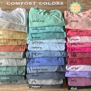May include: Stacks of colorful t-shirts in various shades, including white, ivory, and peachy. Each shirt is labeled with its color name, such as "island reef" and "crunch berry". The shirts are neatly folded, showcasing a range of comfort colors.