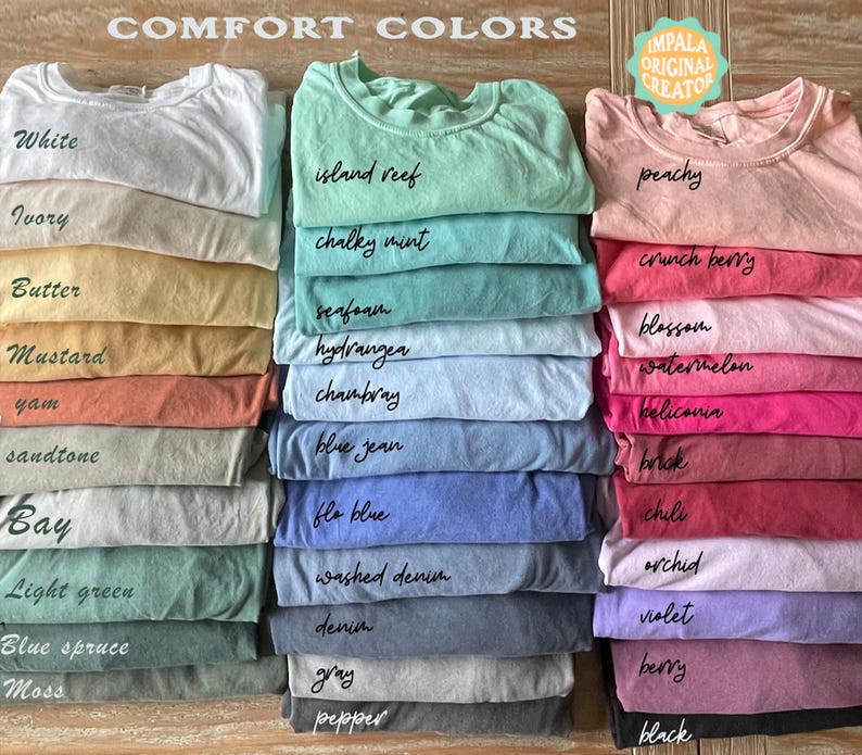 May include: Stacks of colorful t-shirts in various shades, including white, ivory, and peachy. Each shirt is labeled with its color name, such as "island reef" and "crunch berry". The shirts are neatly folded, showcasing a range of comfort colors.