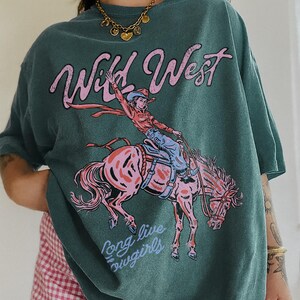 Puede incluir: Una camiseta de manga corta color verde azulado oscuro con un estampado gr&aacute;fico de una persona montando a caballo. El texto "Wild West" aparece en rosa sobre el jinete, y "Long live cowgirls" est&aacute; impreso en azul claro bajo el caballo.