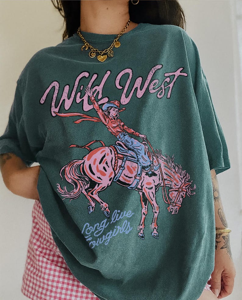 Puede incluir: Una camiseta de manga corta color verde azulado oscuro con un estampado gr&aacute;fico de una persona montando a caballo. El texto "Wild West" aparece en rosa sobre el jinete, y "Long live cowgirls" est&aacute; impreso en azul claro bajo el caballo.