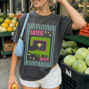 May include: Dark gray t-shirt with a graphic of a green alligator and the words "LATER GATOR" in pink and white. The shirt is paired with white shorts and a light blue purse. The background includes produce stands.