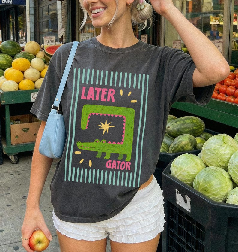 May include: Dark gray t-shirt with a graphic of a green alligator and the words "LATER GATOR" in pink and white. The shirt is paired with white shorts and a light blue purse. The background includes produce stands.