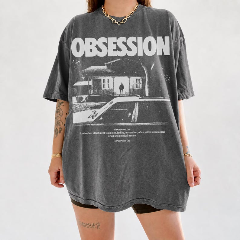 May include: A charcoal grey oversized t-shirt featuring the word OBSESSION in large white letters. Below is a black and white graphic of a house and a car, with dictionary-style text defining obsession as a relentless attachment to an idea.