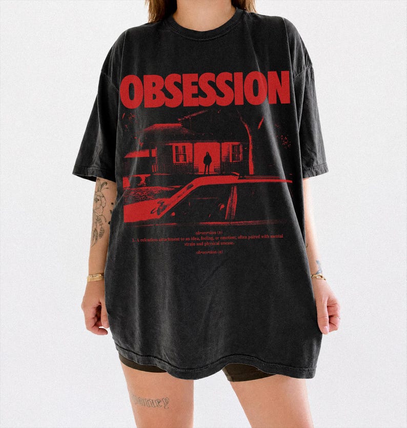 May include: A black oversized t-shirt featuring the word OBSESSION in bold red capital letters. Below the text is a red graphic illustration of a house and a car. Small red text defines the word obsession, including the phonetic pronunciation.