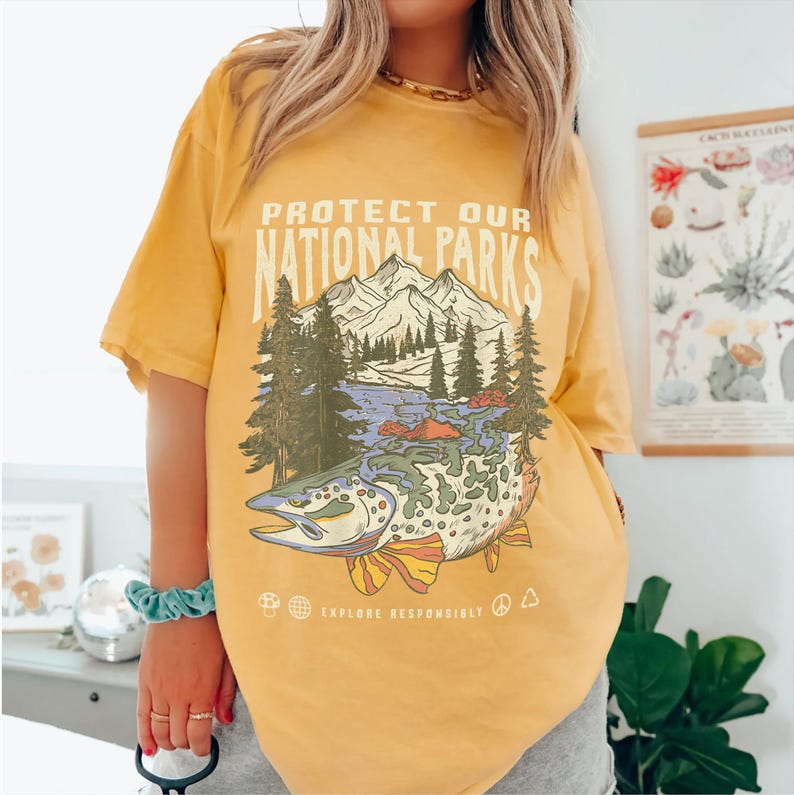 May include: Yellow short-sleeved t-shirt with a graphic print of a fish, mountains, and trees. The text "Protect Our National Parks" is printed on the shirt. The text "Explore Responsibly" is printed below the graphic.