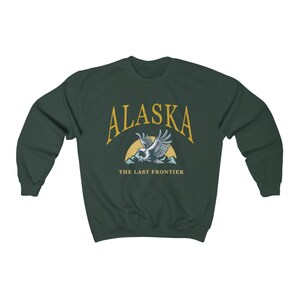 Peut inclure: Un sweat-shirt vert fonc&eacute; avec le texte "Alaska" en jaune, un graphique d'un aigle volant au-dessus des montagnes et le texte "The Last Frontier" en jaune.