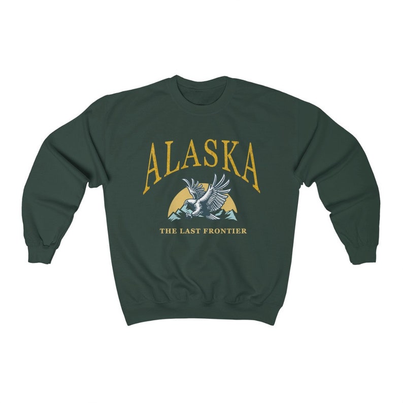 Peut inclure: Un sweat-shirt vert fonc&eacute; avec le texte "Alaska" en jaune, un graphique d'un aigle volant au-dessus des montagnes et le texte "The Last Frontier" en jaune.